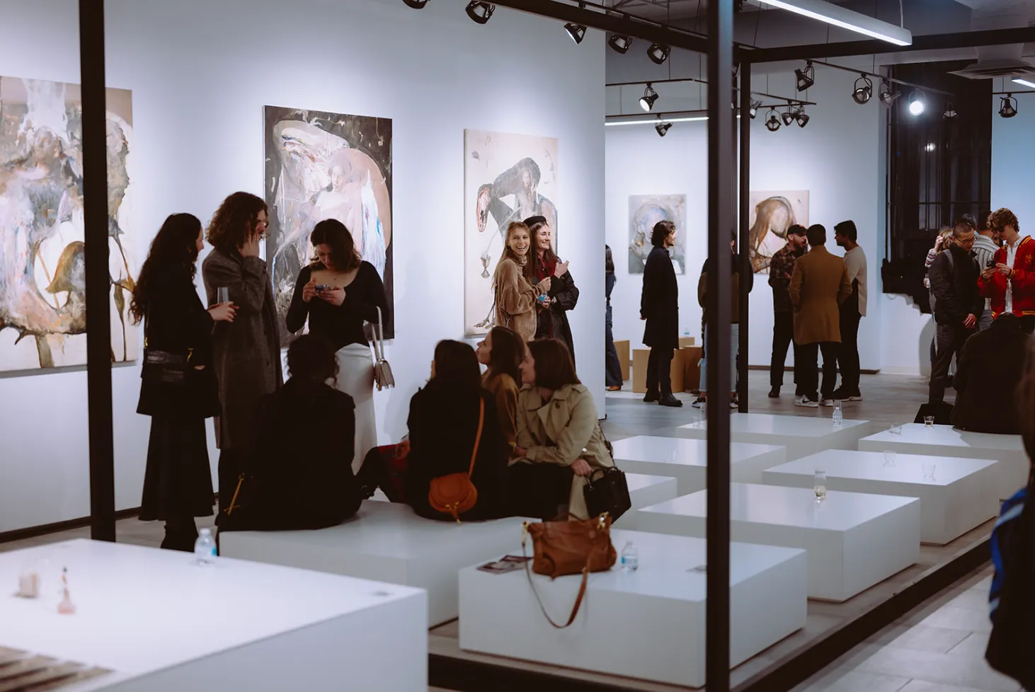 People socializing and viewing abstract paintings in a modern art gallery with white walls and minimalist seating.